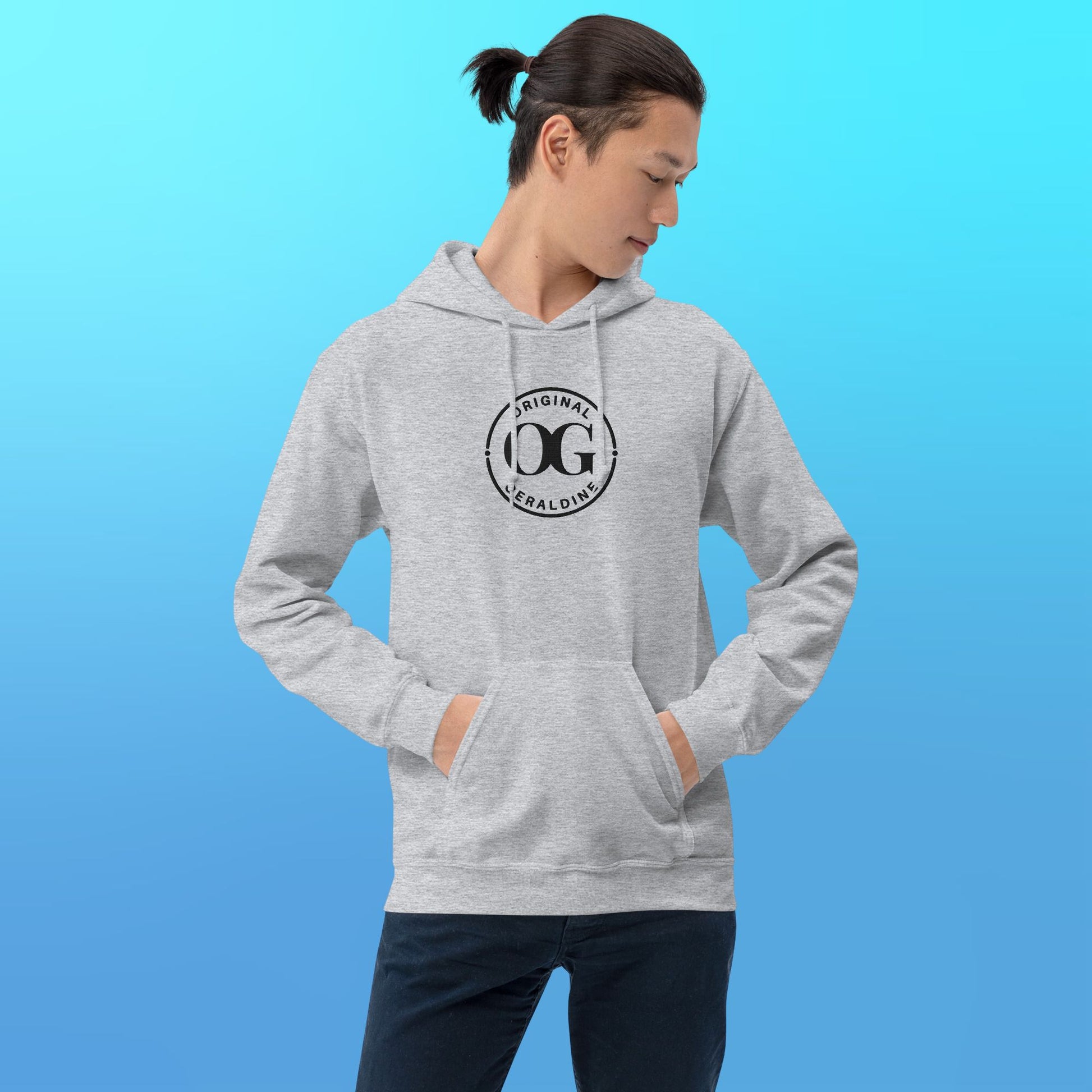 A person wearing a heather grey pullover hoodie with front pocket pouch with OG Original Geraldine emroidered in black thread on the center front.