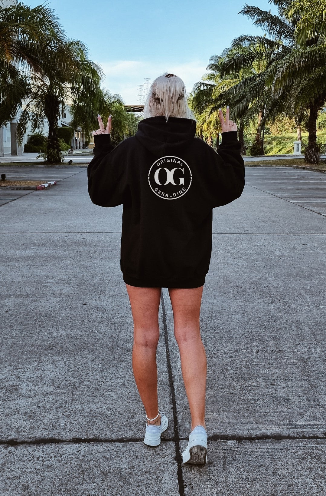 A woman showing the back of a black zip up hoodie with OG Original Geraldine printed on the upper back area in a circle.