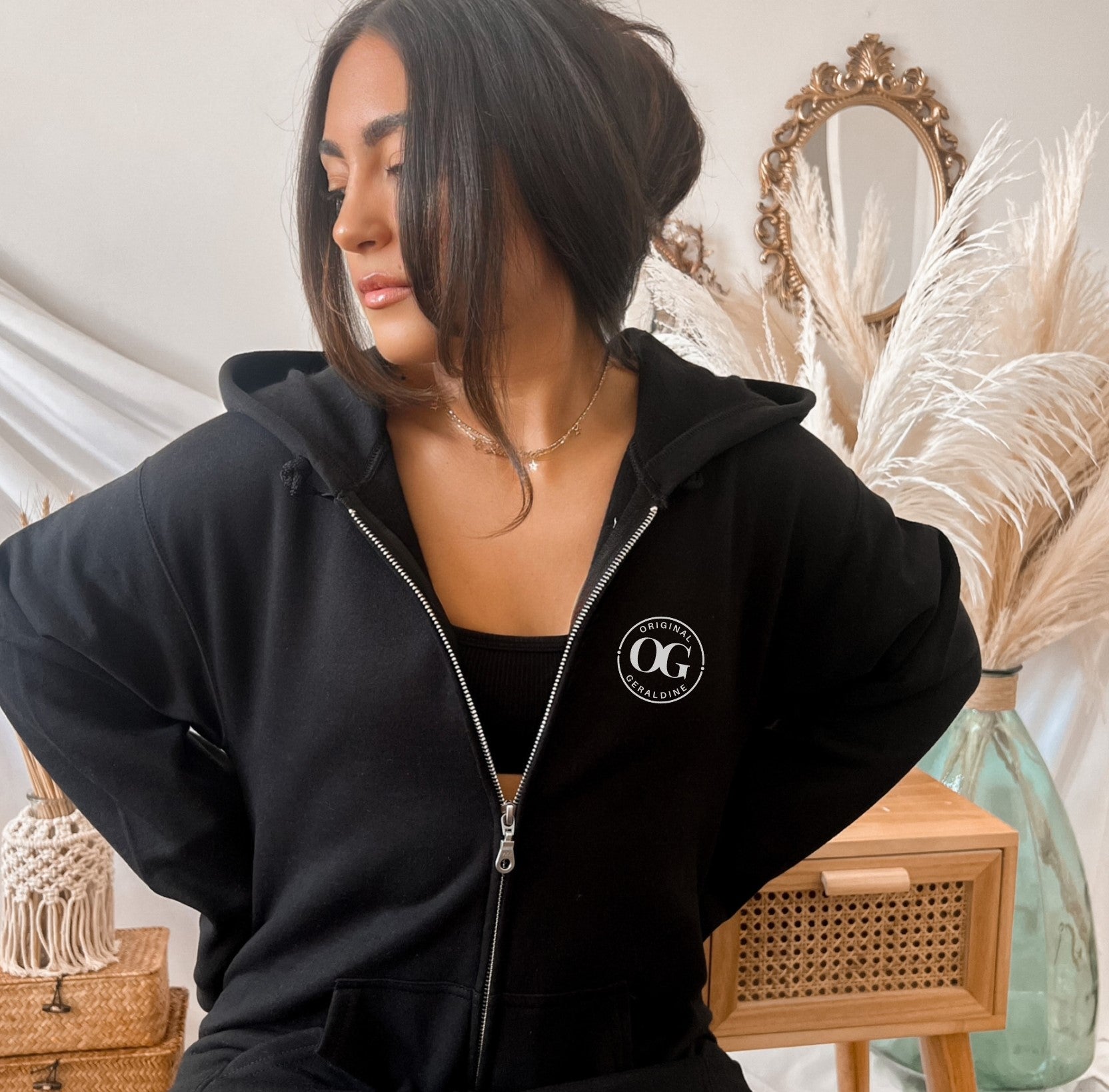 A woman wearing a black zip up hoodie with OG Original Geraldine printed on the left upper chest area in a circle.