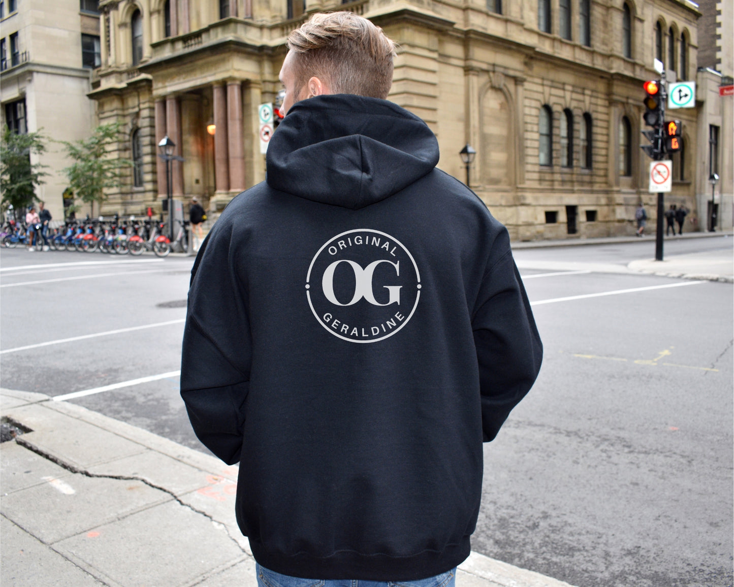 A woman showing the back of a black zip up hoodie with OG Original Geraldine printed on the upper back area in a circle.