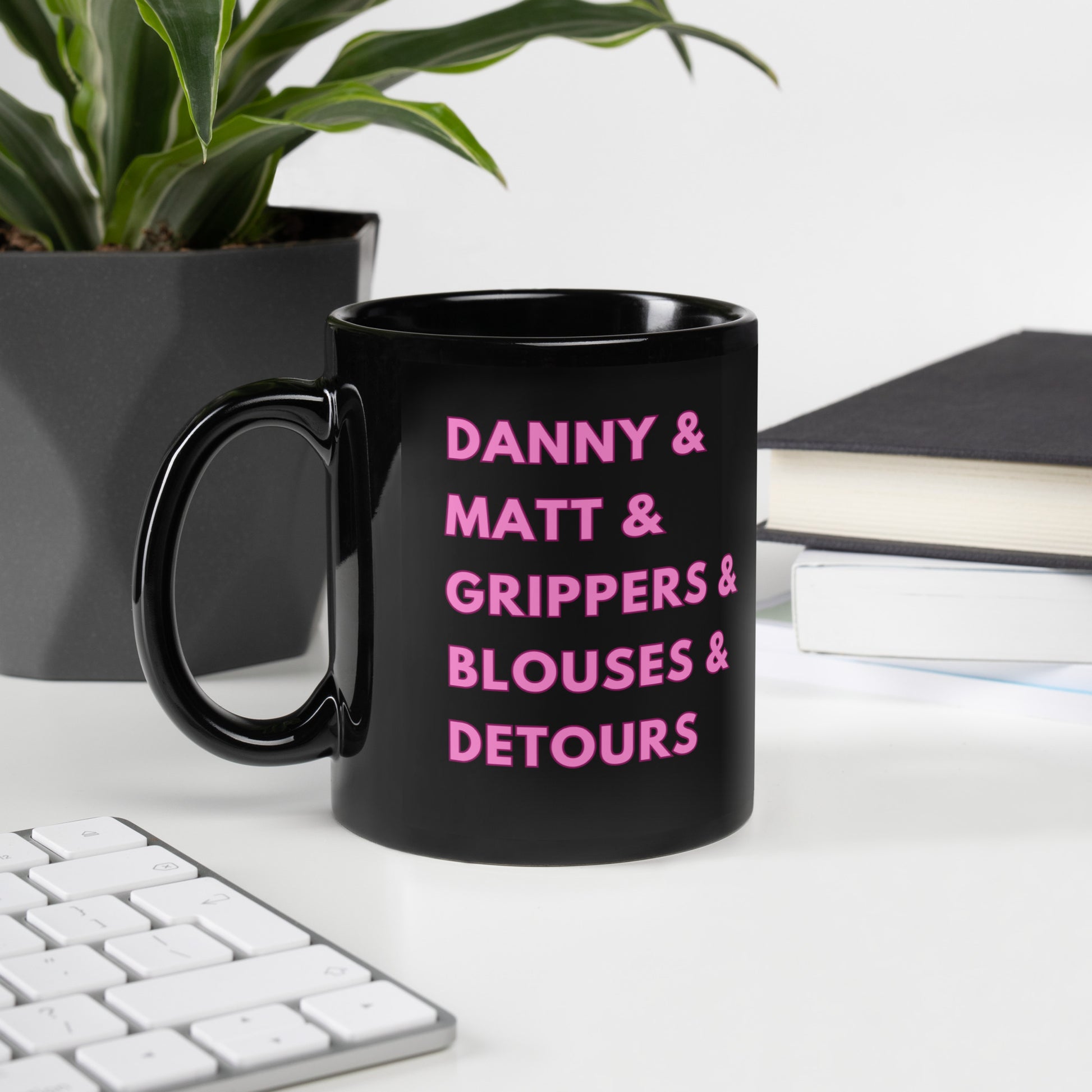 A glossy black mug on an office background that reads "Danny & Matt & Grippers & Blouses & Detours" in pink uppercase font with a darker pink border.