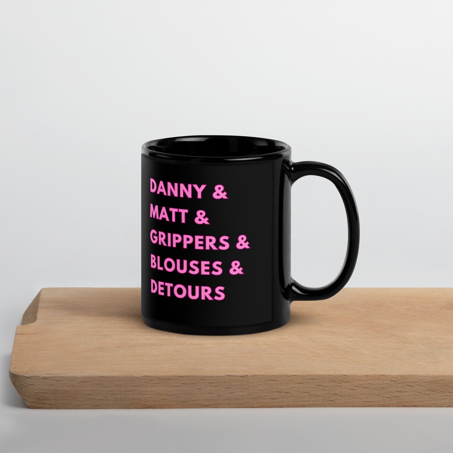 A glossy black mug on a piece of wood with a white background that reads "Danny & Matt & Grippers & Blouses & Detours" in pink uppercase font with a darker pink border.