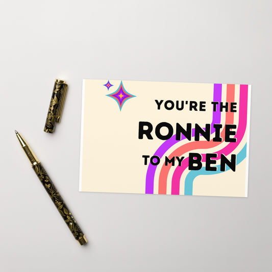 The front of a greeting card that says "You're the Ronnie to my Ben" in black font on top of colorful (blue, pink, orange and purple) curved stripes with two coordinating atomic stars in the upper left corner.