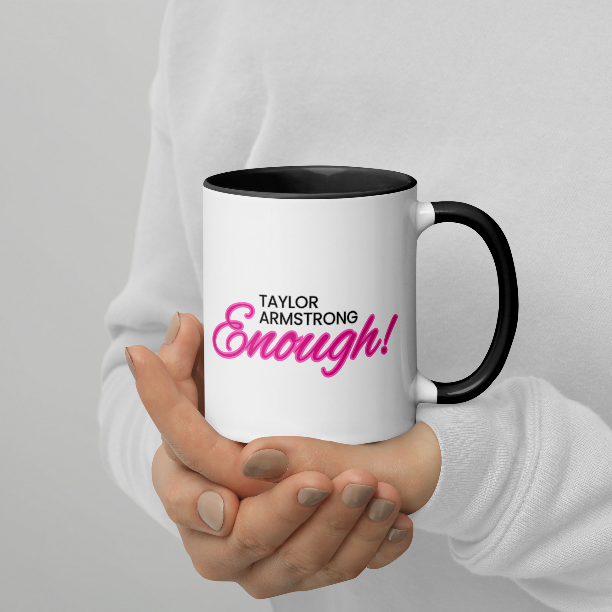 A person holding a white mug with a black handle and black interior with the phrase Taylor Armstrong ENOUGH! written in black and pink font in the center.